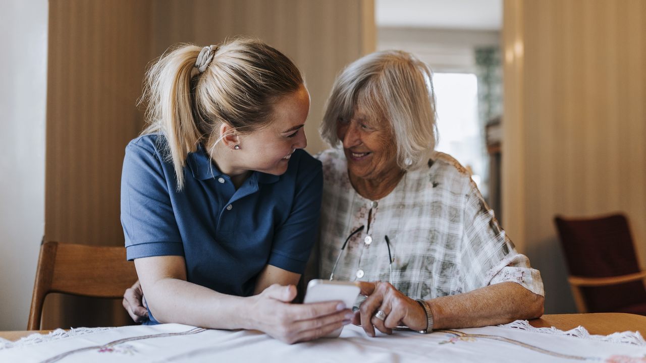 A caregiver smiles at an older woman while they look at the caregiver&#039;s phone together at the kitchen table.