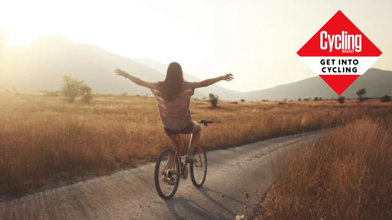 How to learn to ride a bike image is a woman on a bike with her arms out stretched to the sides on a road with brown grasses either side and hills in the background. 