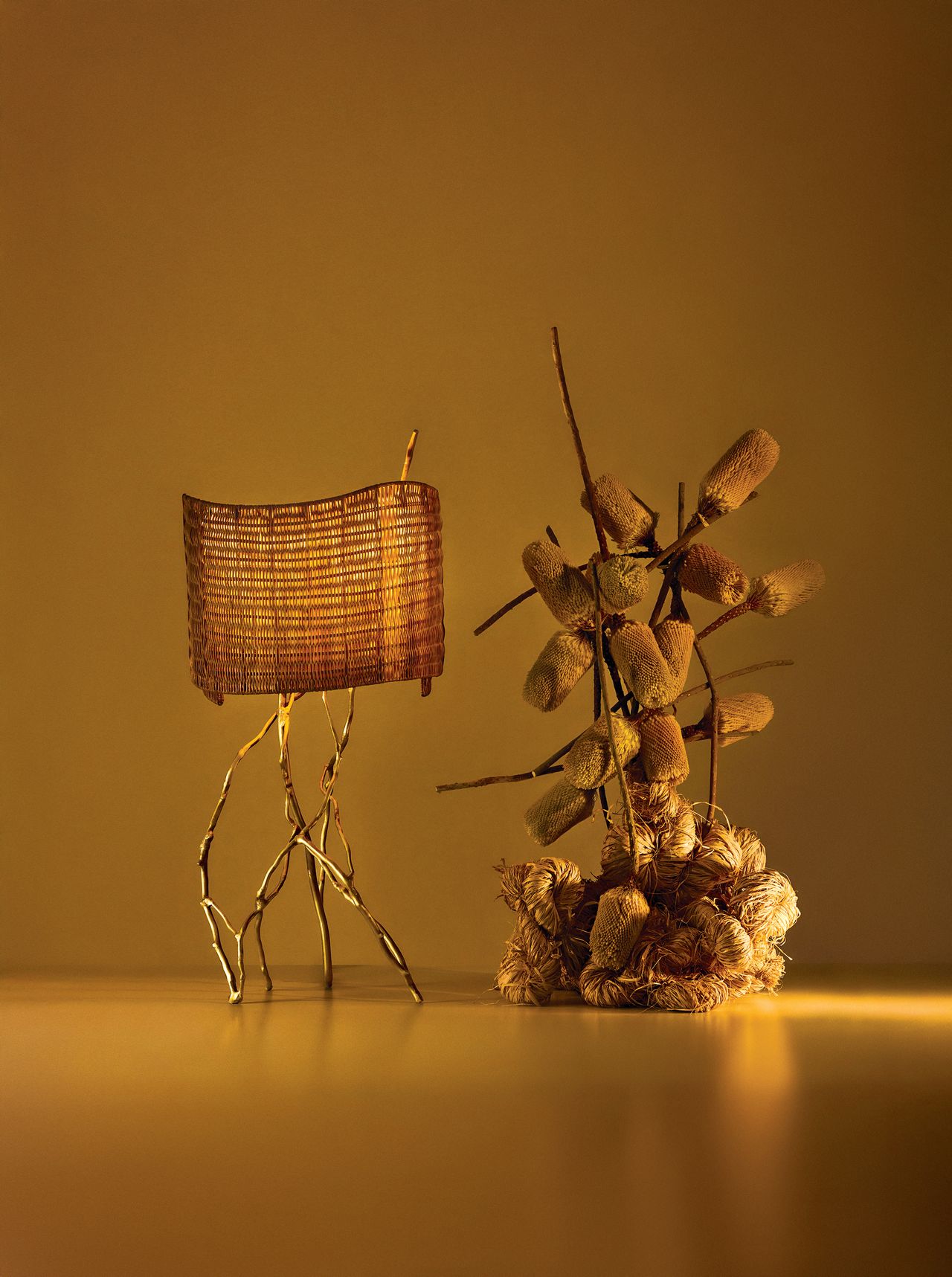 Image of an artisanal lamp next to an artisanal foliage structure in a display setup that is drenched in ochre yellow/gold