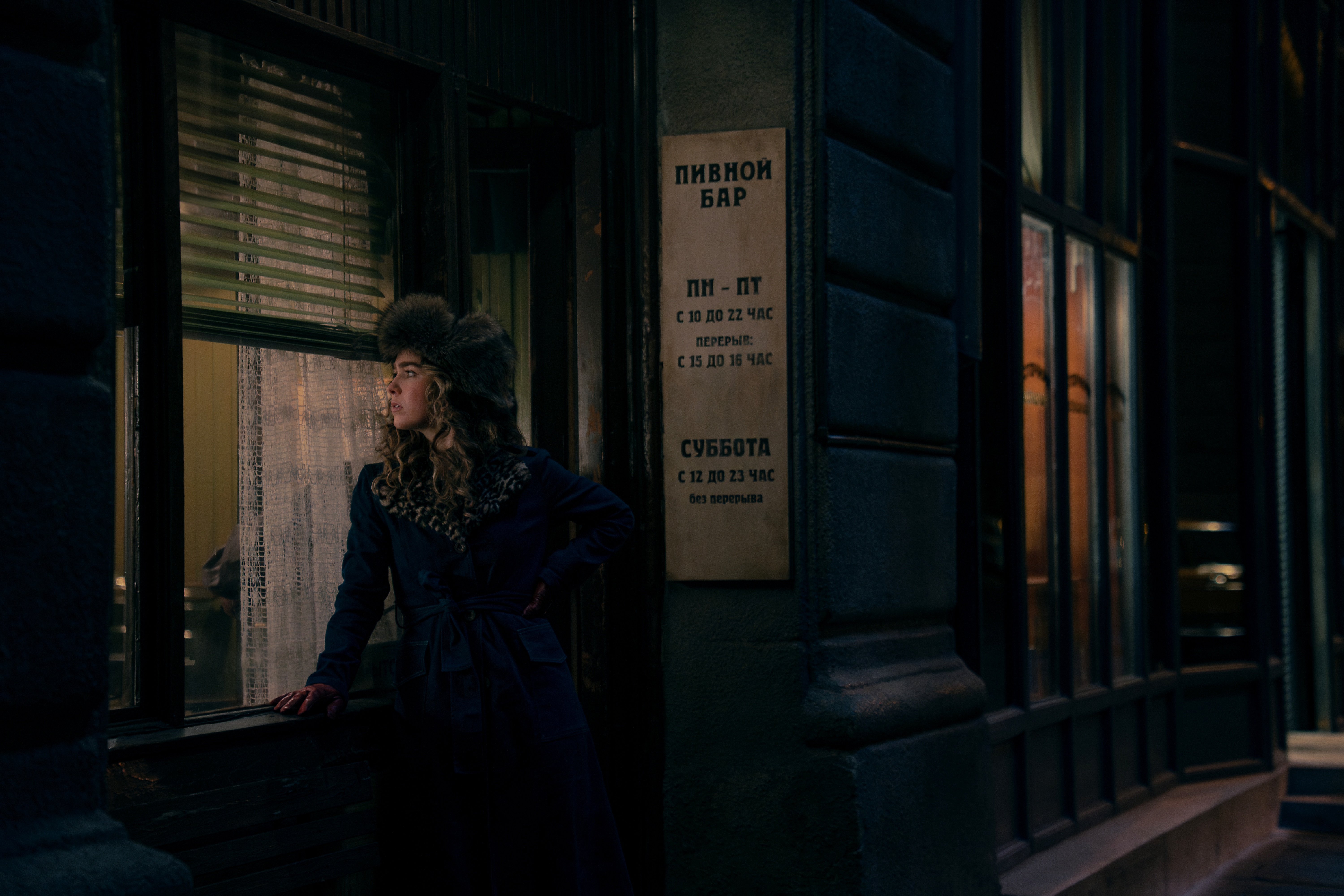 A young woman dressed in a fur leather coat and hat peeks inside a dimly lit pub with a Russian, yellowed sign from a road at night.