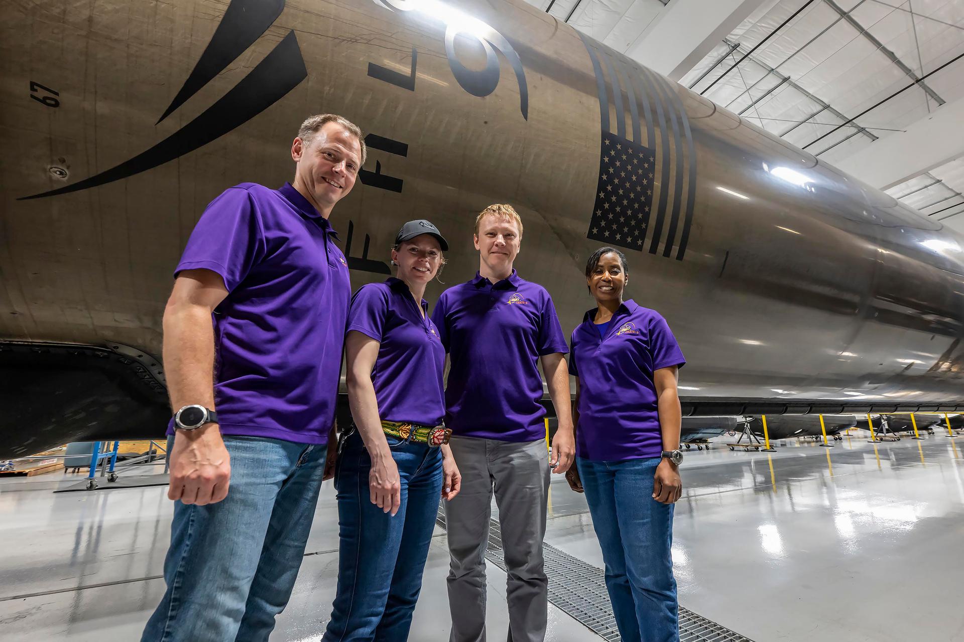 four people in jeans and blue three-button shirts smile beside a giant metal cylinder in a hangar