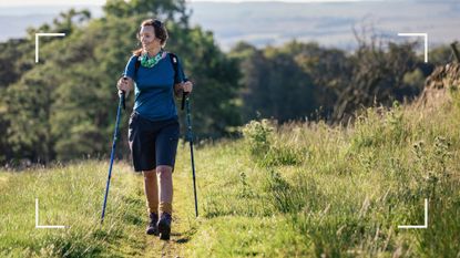 Woman Nordic walking across grassy hill with trees behind her, wearing active clothes and carrying walking poles, smiling
