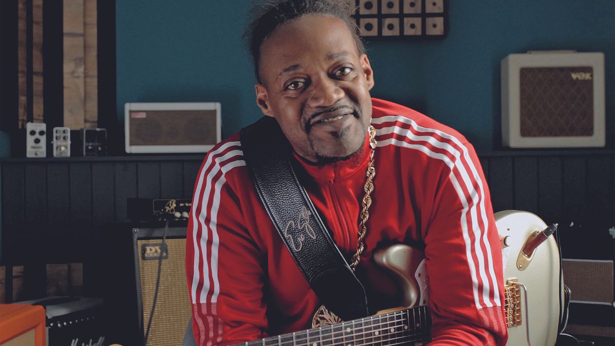 Eric Gales poses in a studio wearing a red track suit and holding his signature Magneto RawDawg electric guitar