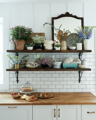 open shelving with ceramicware flowers and a mirror above white kitchen cabinet with wooden worktop
