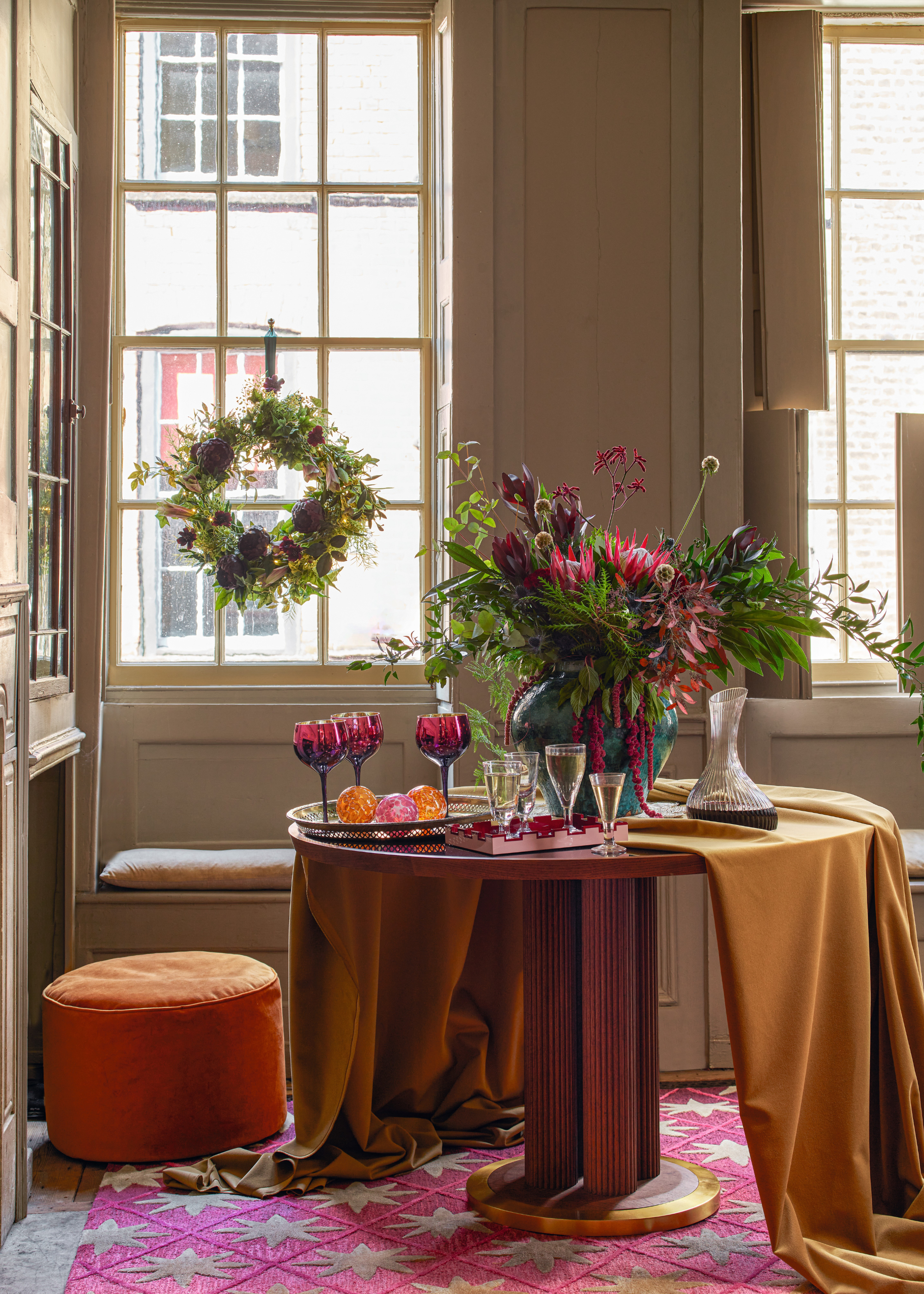 A dining room decorated for Christmas with a long tablecloth draped over half of the table with a large vase of vibrant foliage in the center of the table and a wreath hanging in the middle of the window