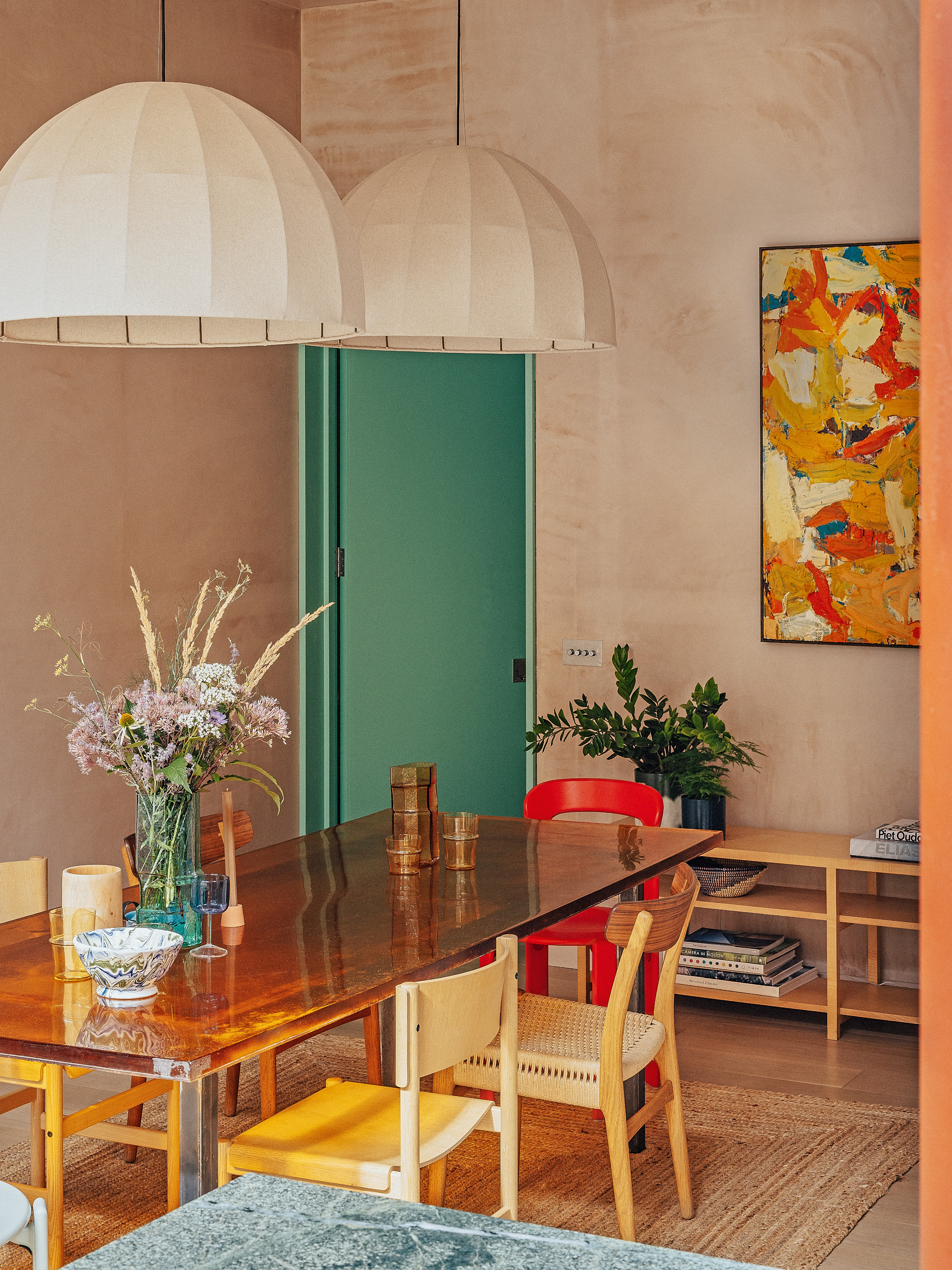 Dining room with plaster pink walls, amber glass and steel dining table, mixed wood and cane chairs, oversized natural pendant lights, jute floor rug and red and yellow abstract art