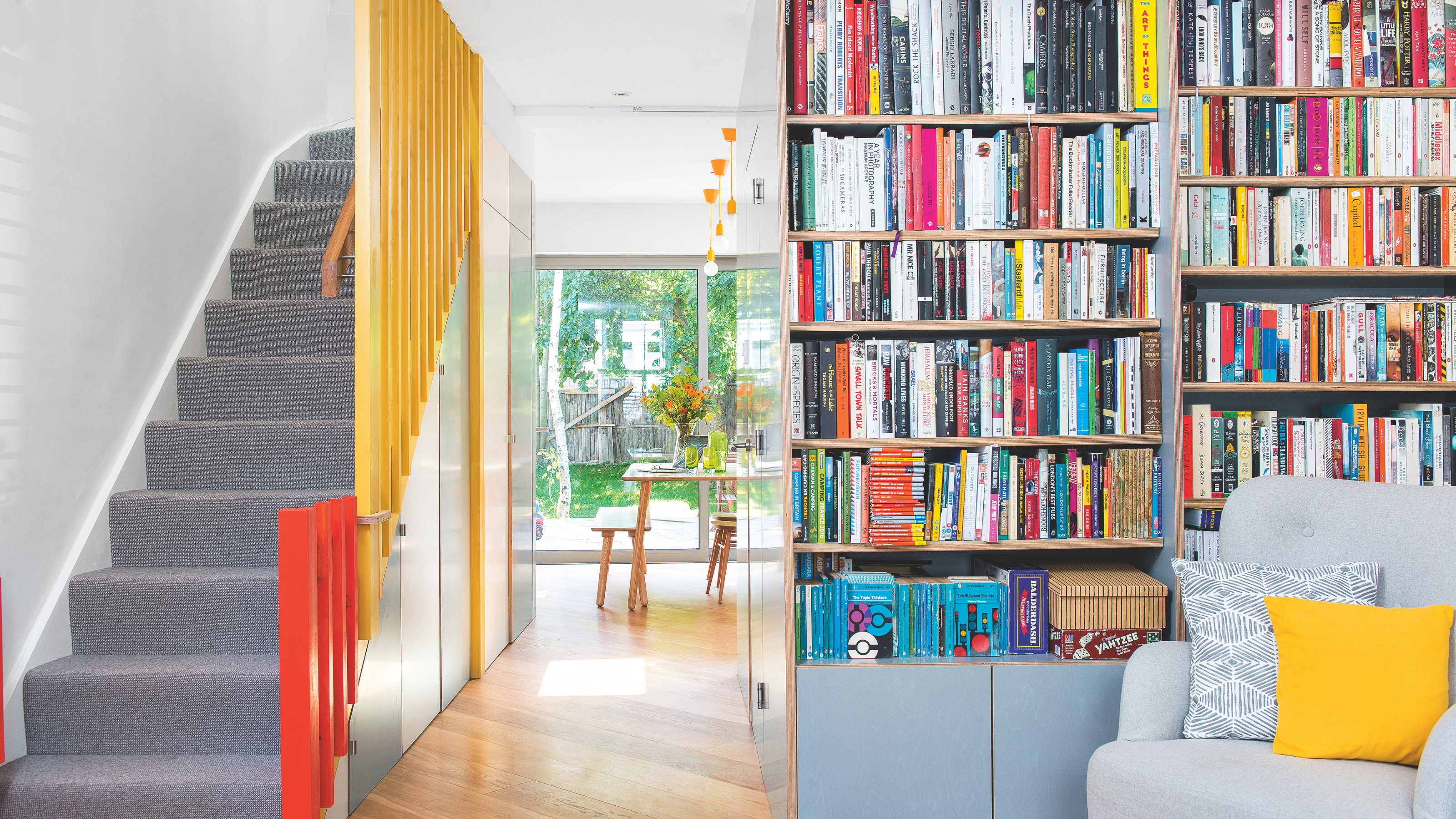 Hallway with storage and bookcase and doors painted in duck egg blue, with staircase opposite with grey stair carpet and yellow and orange painted bannisters.