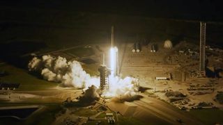 aerial view of a rocket lifting off into the night sky, its bright white thrust lighting up its launch pad