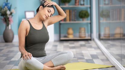 woman sitting crossed legged on a yellow mat on a grey lino floor tilting her head to the side and holding it with one hand, eyes closed