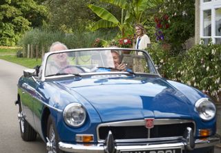 Richard (Peter Davison) and Anne (Barbara Flynn) drive away from the cottage in a blue open-topped classic car. Martha (Sally Bretton) is standing at the gate, apprehensively watching them leave.