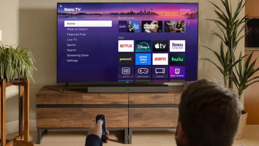 A person seated in the foreground of a living room browses the Roku home screen on a TV that sits in the background