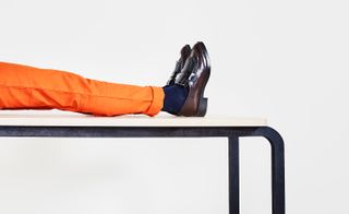 pair of legs on a white table top wearing orange pants, black socks & brown shoes