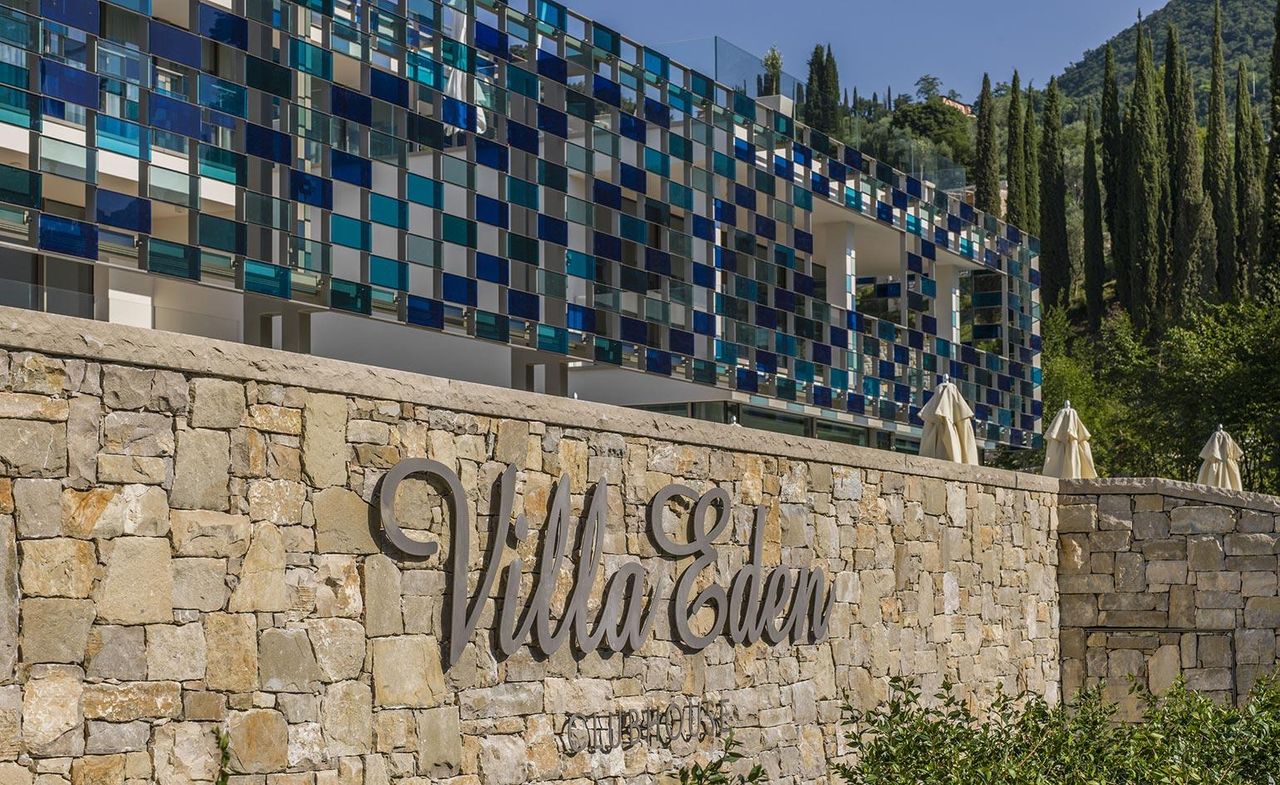 Exterior view of the Villa Eden. The top part is a glass exterior in different shades of blue. The bottom part is stacked rocks with the writing &quot;Villa Eden&quot;. We see a mountain to the right.