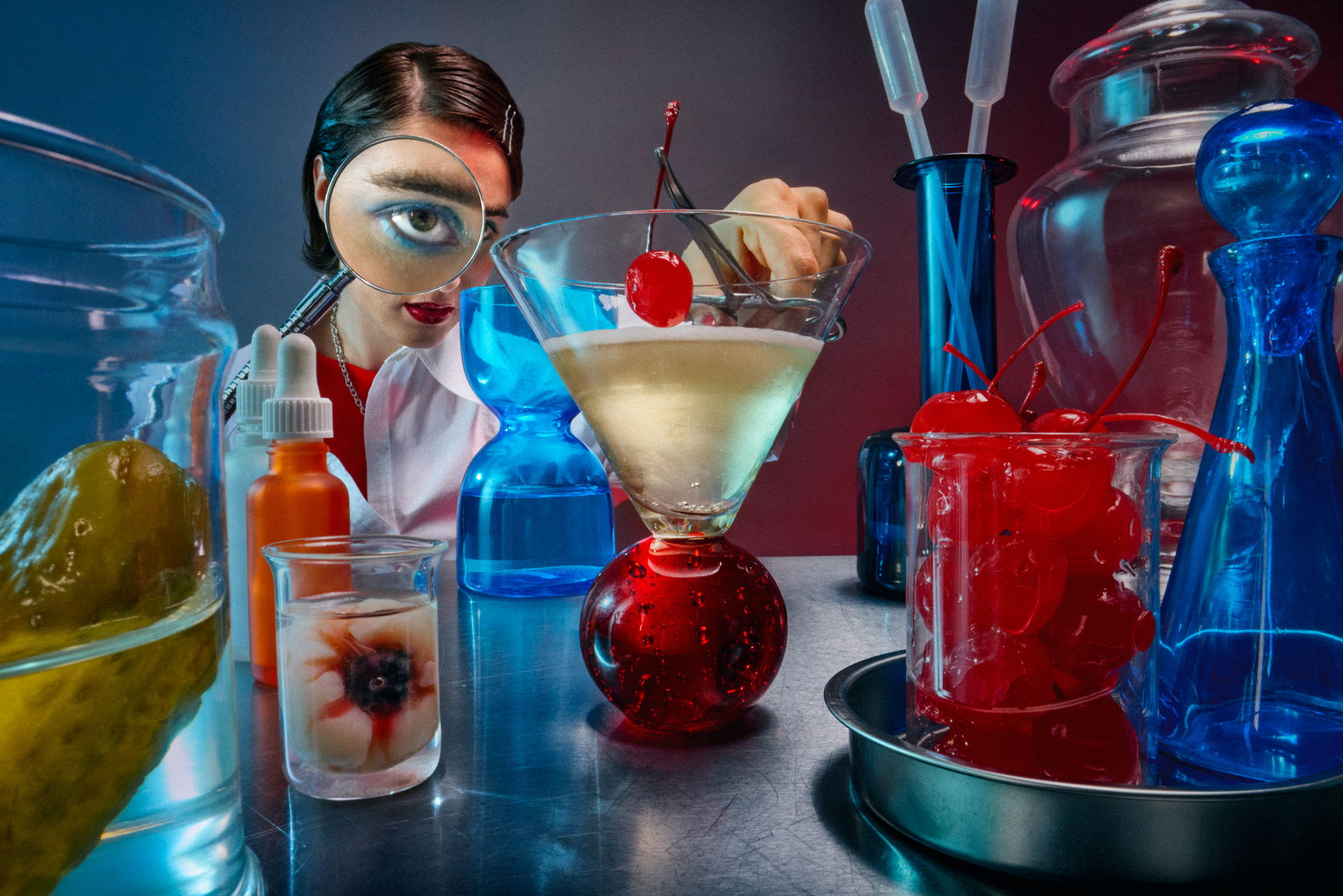 A person in a lab coat peers through a magnifying glass at a cocktail topped with a cherry, surrounded by colorful jars and glassware on a reflective surface