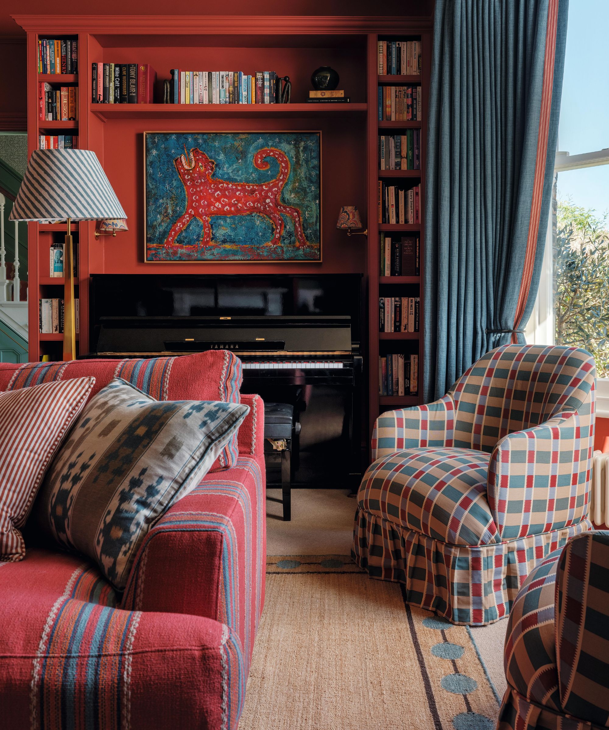 A red painted sitting room with large window dressed with blue drapes, a bespoke bookcase area built around a piano, a striped red sofa and a checked armchair pair