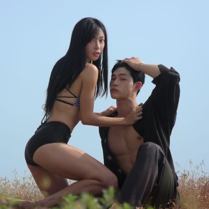 A woman (Kim Min-gee) wears a bikini top and shorts while kneeling and holding the collar of a man (Song Seung-il) wearing a black button-down shirt open with gray pants. The man rests a hand on his head while sitting on the ground, as the pair pose outdoors on a hill. A still from the Korean reality show 'Single's Inferno' season 5.