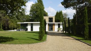 modern house finished in white render and timber cladding with long gravel driveway, lawn and conifer lined small avenue