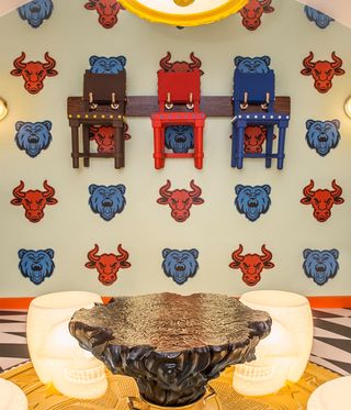 Always Close installation by Studio Job in Luxembourg. A wall with a bull and lion head pattern, chair shaped coat hangers, a tree trunk shaped coffee table in front of it and skull shaped chairs.