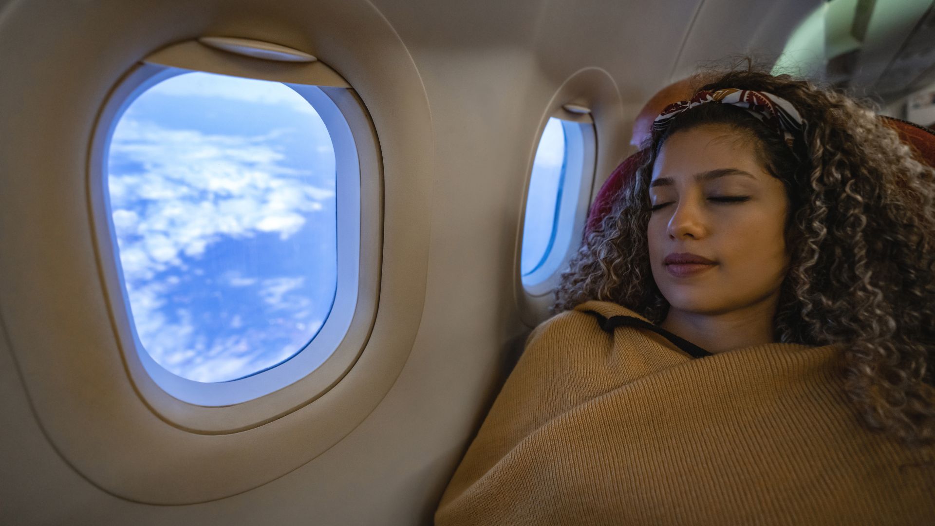 A young woman with curly hair sleeps on the window seat of a plane while draped in a tan blanket.