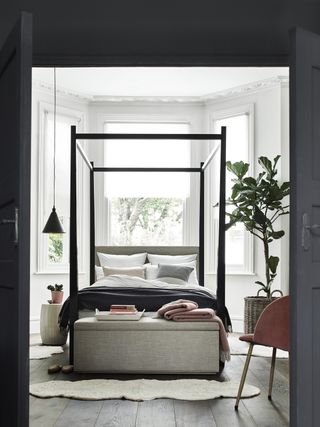 white bedroom with gray bed, grey ottoman, black four poster frame, black pendant, fig tree, armchair, coral accents