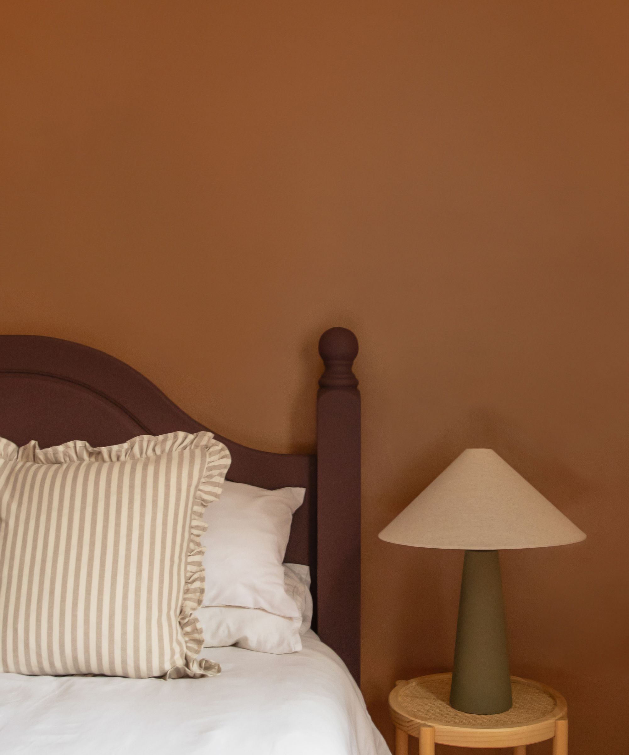 bedroom with brown wall, brown timber bedhead, striped cream and white pillow with ruffle edge, timber side table with lamp styled on it, and white bed linen