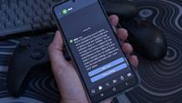 A hand holds a Samsung Galaxy Z Flip 7, showing an Xbox app notification about verified adult account status. Two controllers and a keyboard sit slightly out of focus.