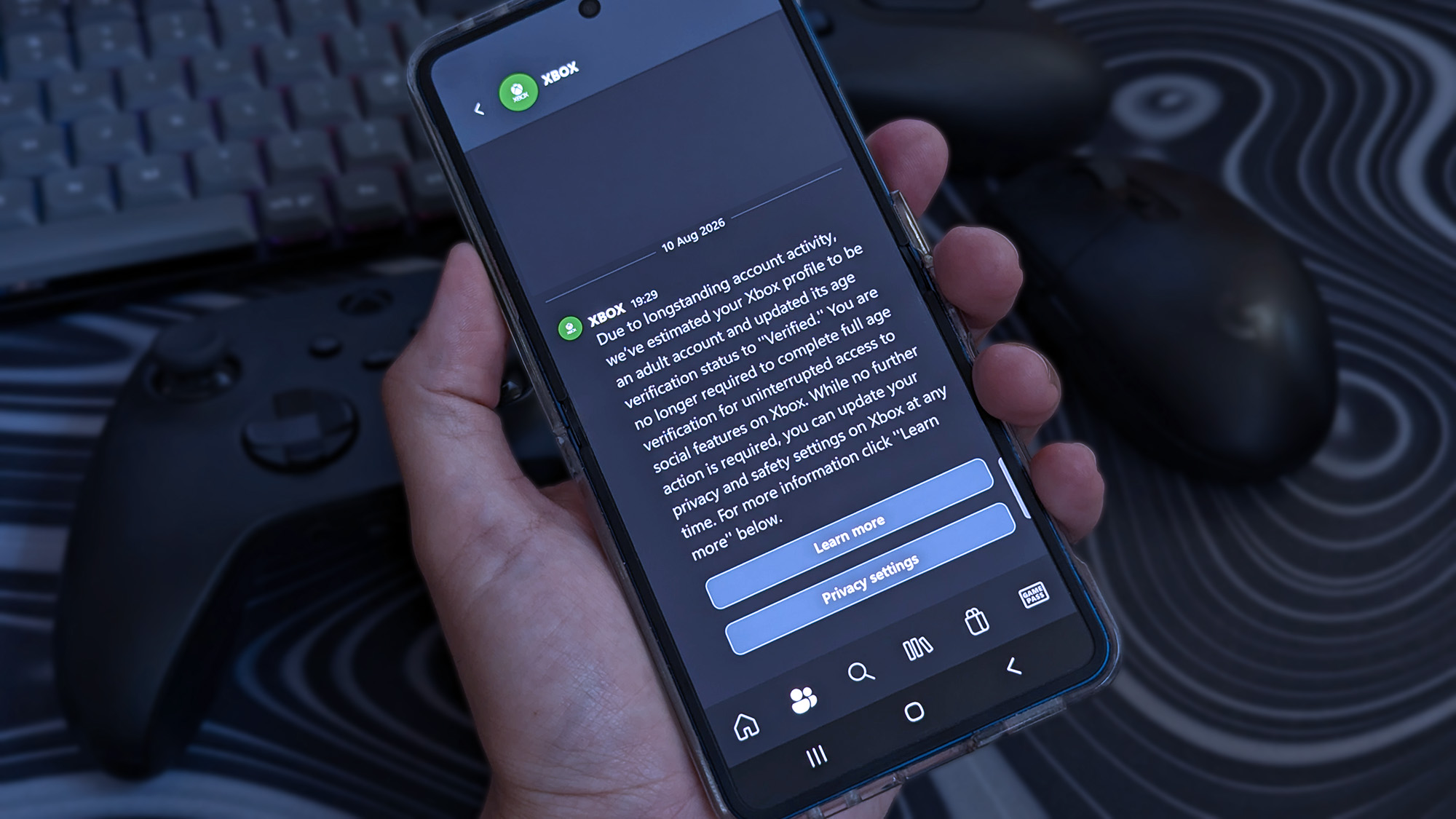 A hand holds a Samsung Galaxy Z Flip 7, showing an Xbox app notification about verified adult account status. Two controllers and a keyboard sit slightly out of focus.