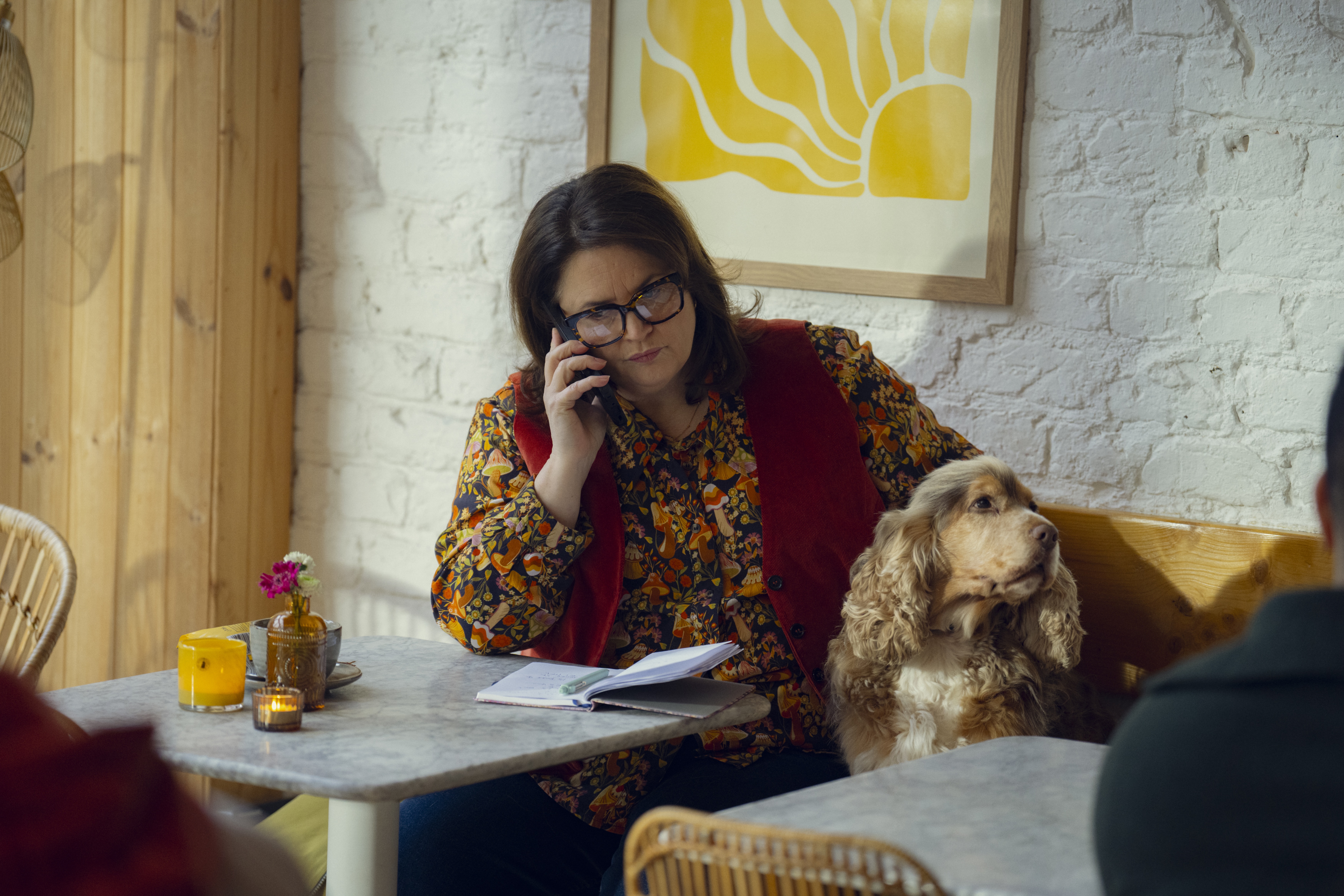 Ruth Jones as Elena Ravenscroft sitting in a cafe talking on the phone with a dog sitting next to her on a bench in a still from the tv show run away