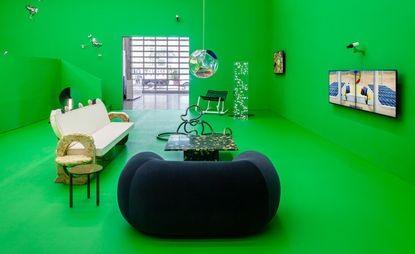 A large lounge area with bright green walls and floor. A dark green velvet armchair sits in the foreground with varying pieces of designer seating around a small dark marble effect coffee table.