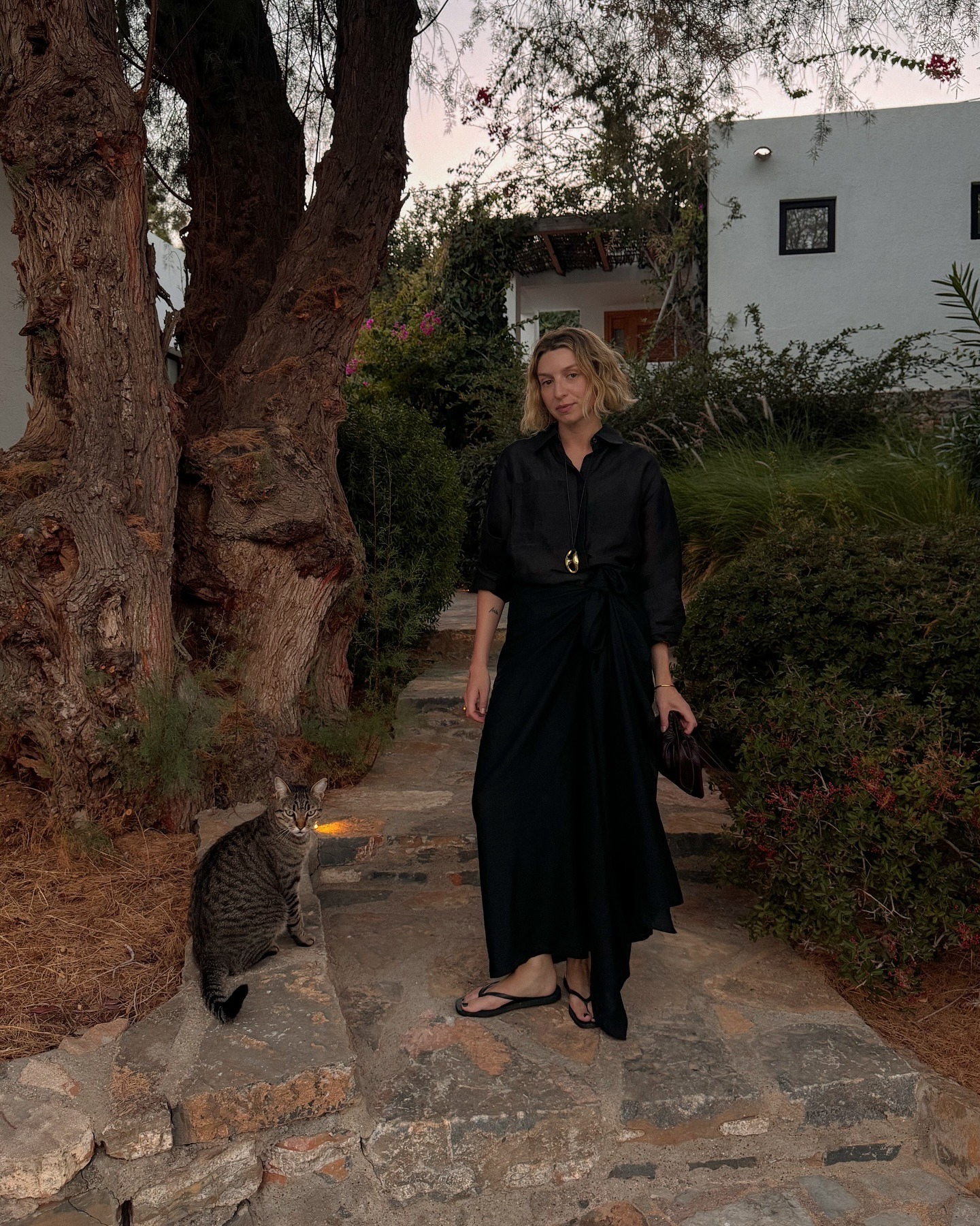 Influencer stands outside on a cobbled pavement wearing a black wrap maxi shirt tucked into a black blouse. She styled her look with black flip-flops and a long corn necklace. A cat sits beside her feet.