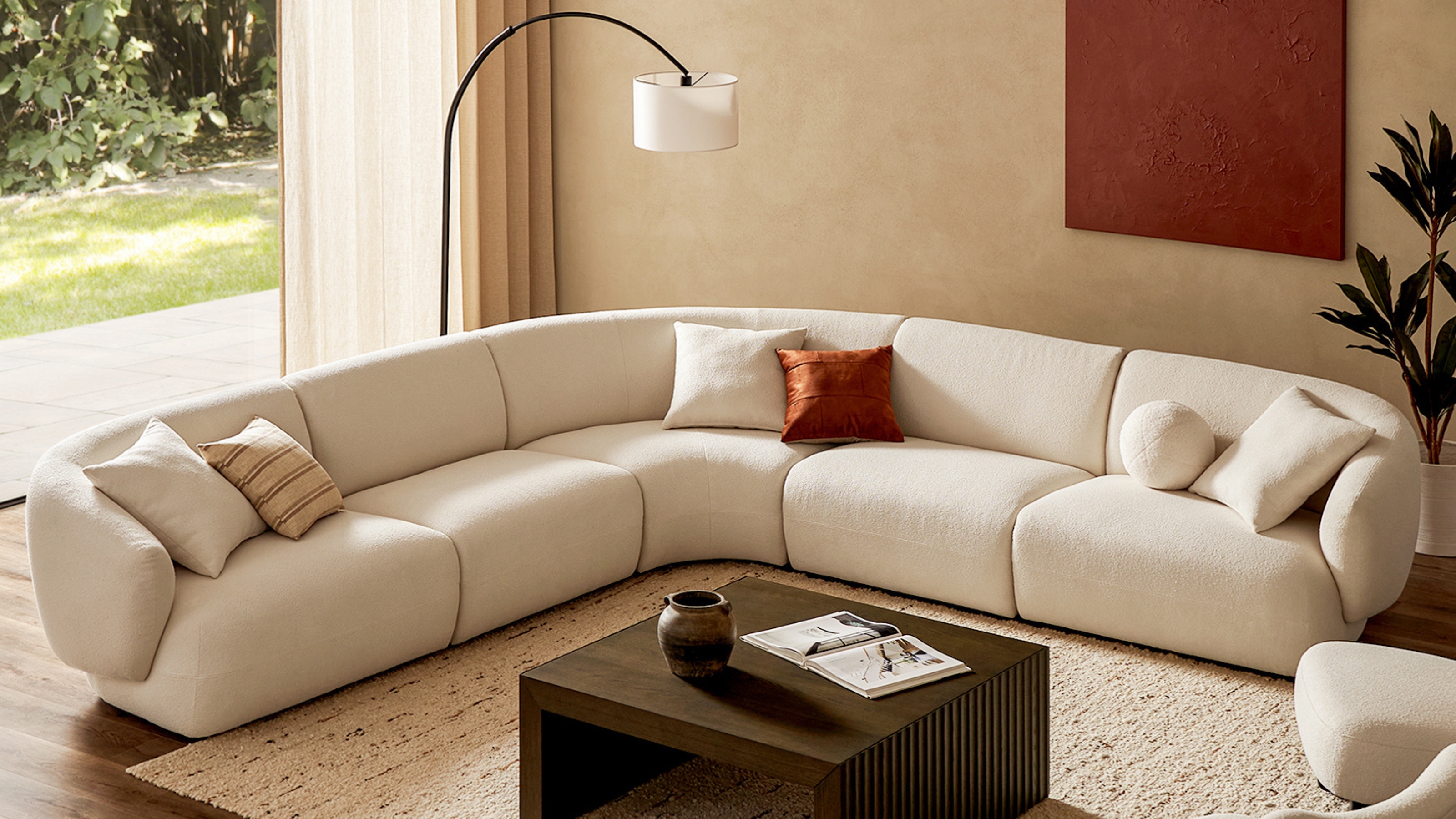 Image of a large, white, L-shaped sectional sofa with six throw pillows in a beige living room. There is a curved floor lamp behind the sofa, and a dark wooden coffee table in the front. 