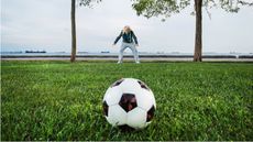 An older man in the distance prepares to run at a soccer ball in the foreground.