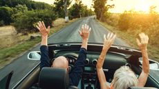 An older man and woman raise their hands in the hair while driving a convertible through the country.