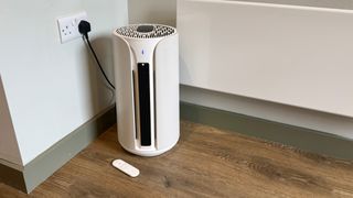 A photo of the Blueair T10i Air Purifier with its remote in a room with white walls and laminated wooden floors