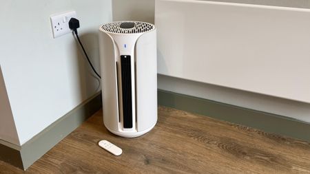 A photo of the Blueair T10i Air Purifier with its remote in a room with white walls and laminated wooden floors