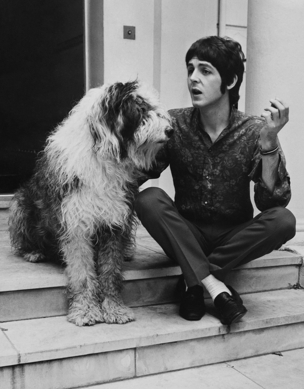Paul McCartney sits on stone steps beside a shaggy old English sheepdog, mid-conversation, in a relaxed black-and-white portrait.