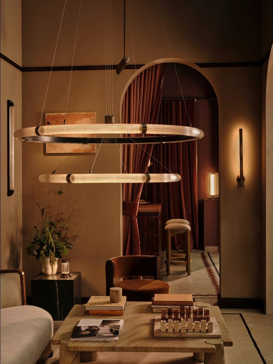 Image of a luxury, beige living room with an arched doorway with a red curtain that leads into a different room. There is a seating area with a square, marble coffee table and a large pendant light hanging over head.