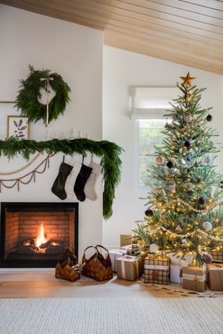 Christmas living room with tree, presents, mantel with foliage and wreath, stockings, open fire, baskets with logs, modern feel