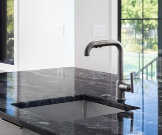 A modern kitchen black stainless faucet detail on an island with a black marble countertop, black frame windows, and white walls