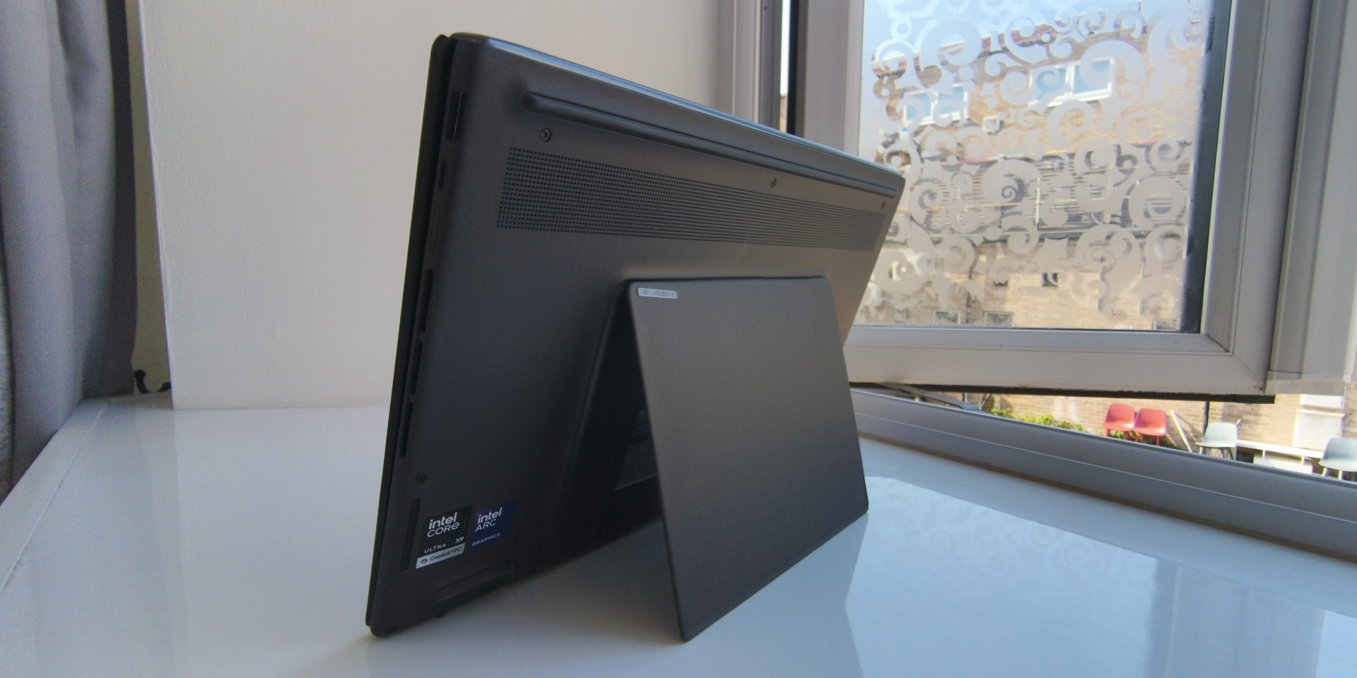 An angled rear view of the dark grey ASUS Zenbook Duo (2026) laptop standing upright on a white surface next to a window, supported by its built-in kickstand plate.