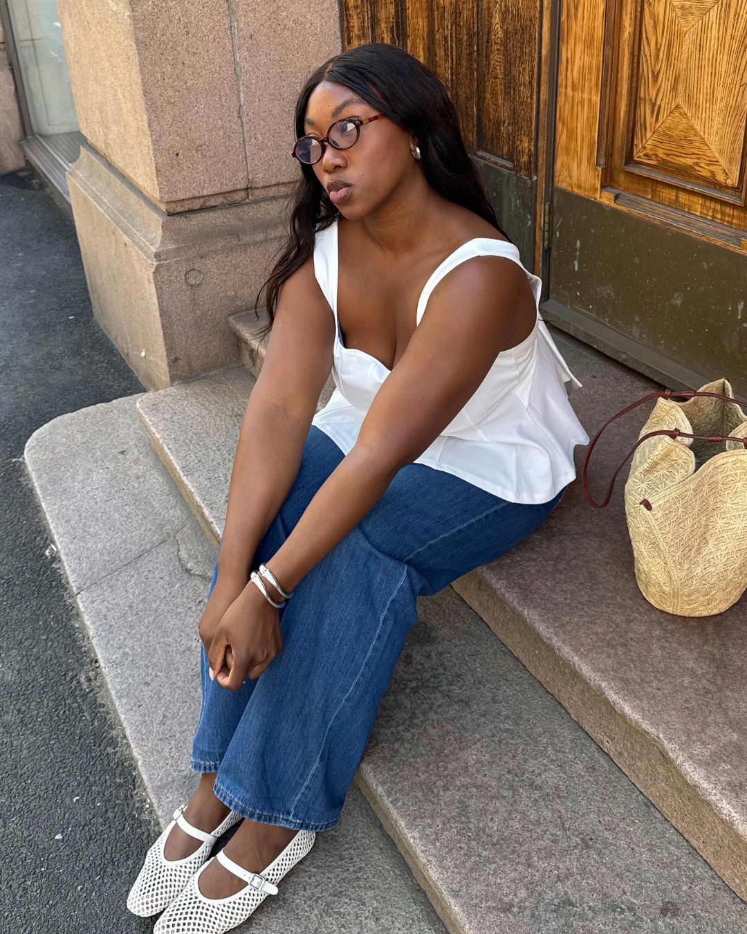 Influencer @nnennaechem sits on some steps outside wearing white mesh ballet flats with a white blouse, jeans and a wicker bag.