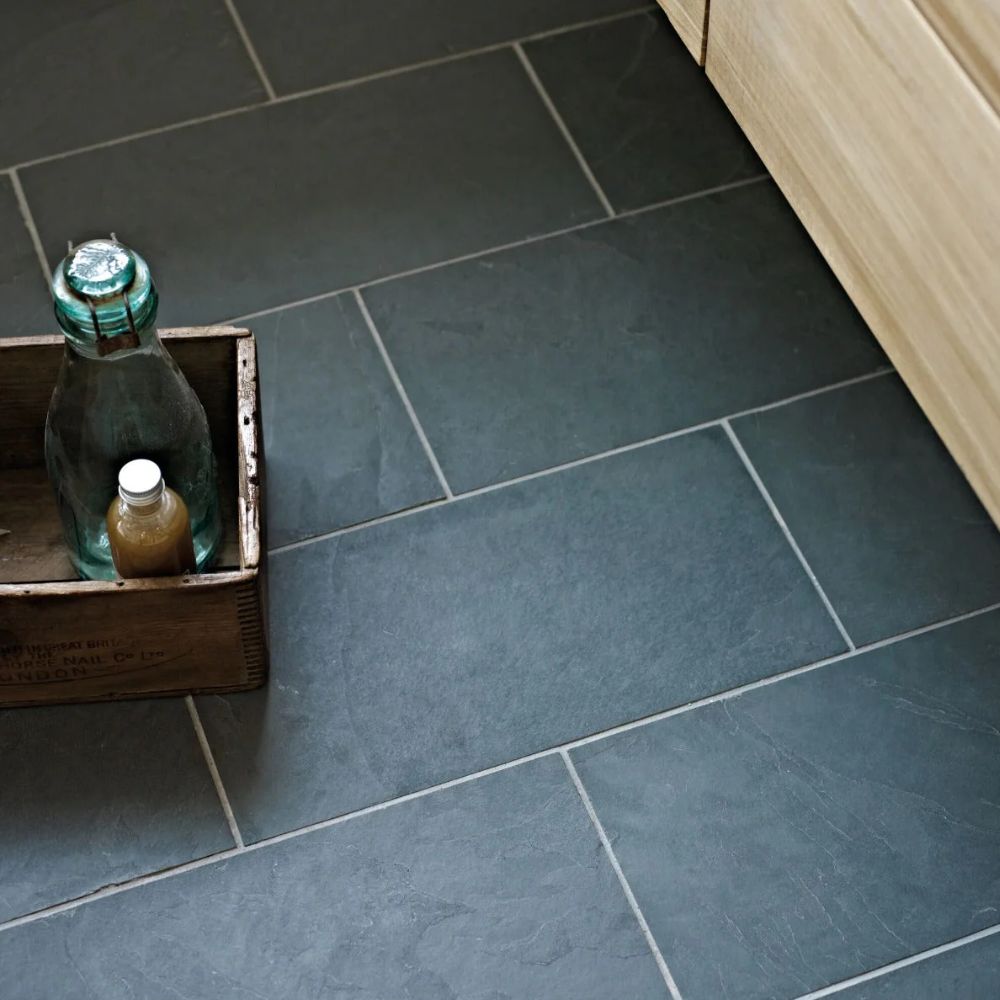 A close-up of dark grey-green slate floor tiles in a mixed rectangular format laid in a staggered pattern, beside a light oak skirting board. A rustic wooden crate holding a glass bottle and small toiletry bottle sits on the textured stone surface.