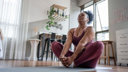 Woman doing reformer Pilates at home with yoga mat, sitting in the middle of her living room stretching with eyes closed, peaceful expression