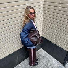 Torivan wears blue bomber and brown athletic pants with a pink stripe while holding a large brown leather tote