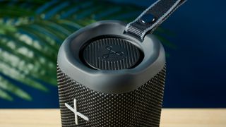 a close up picture of the woofer of a black oblong-shaped bluetooth speaker with mesh covering and a carry strap photographed on a beige table in front of a navy wall