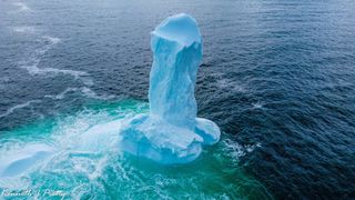 A penis-shaped iceberg floats in the water with two icy ball-shaped structures at its base.