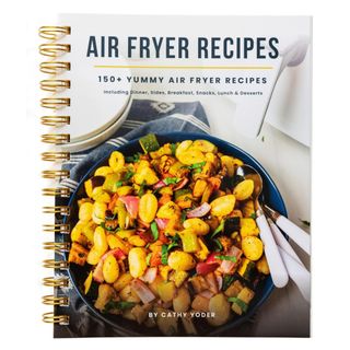 A spiral bound air fryer recipe book with a large image of gnocchi with mixed veg in a blue bowl with pink cutlery on top of a blue patterned tea towel with two white stacked bowls