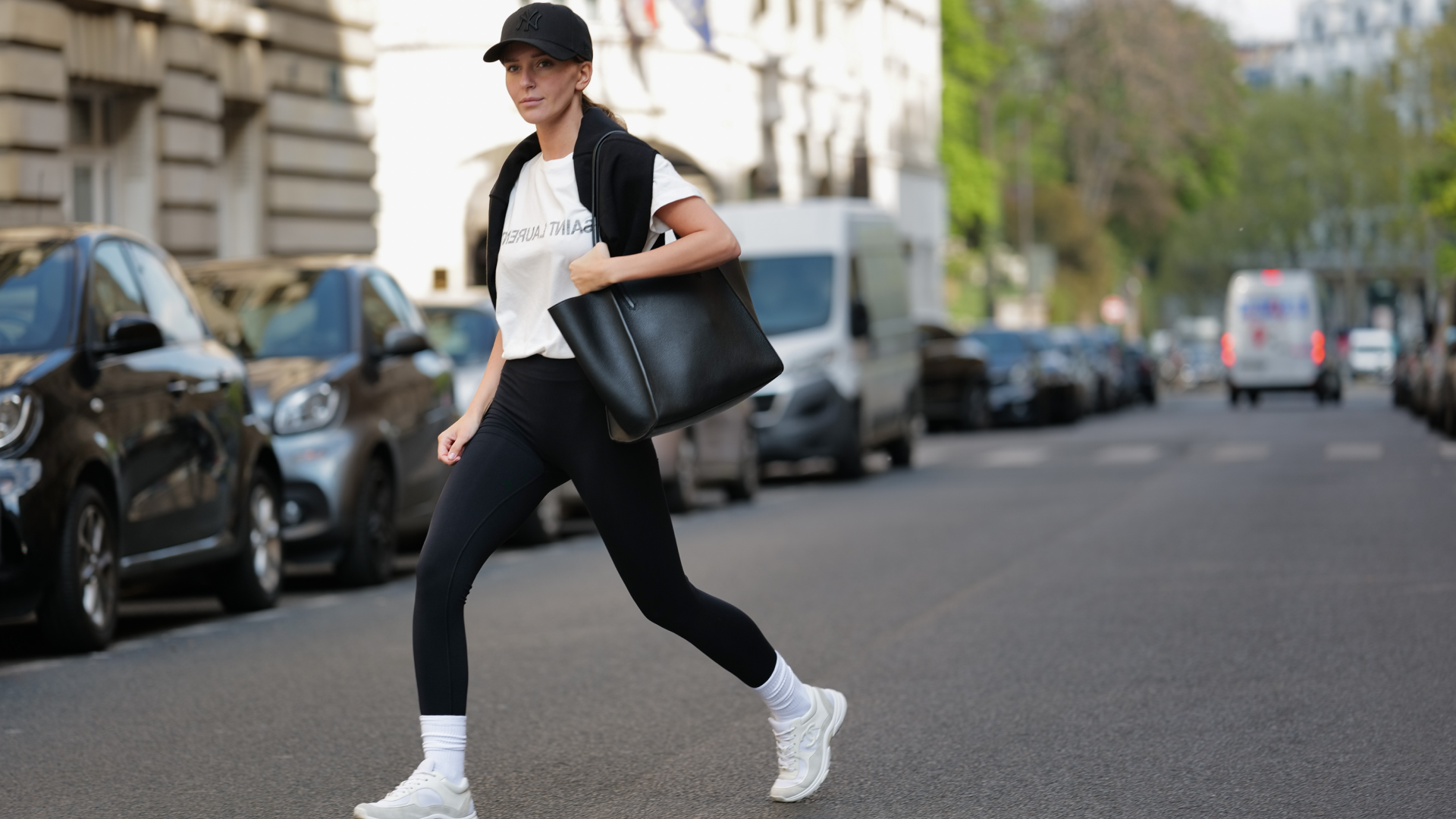 Diane Batoukina wears a black with embroidered NY logo pattern cap from New Era, a white with black faded logo print pattern t-shirt from Saint Laurent Paris, a black wool pullover knot at the shoulder, a black shiny leather large shoulder bag from Zadig&Voltaire, black legging pants, white socks, white leather and suede sneakers from Chanel, during a street style fashion photo session, on April 21, 2023 in Paris, France.