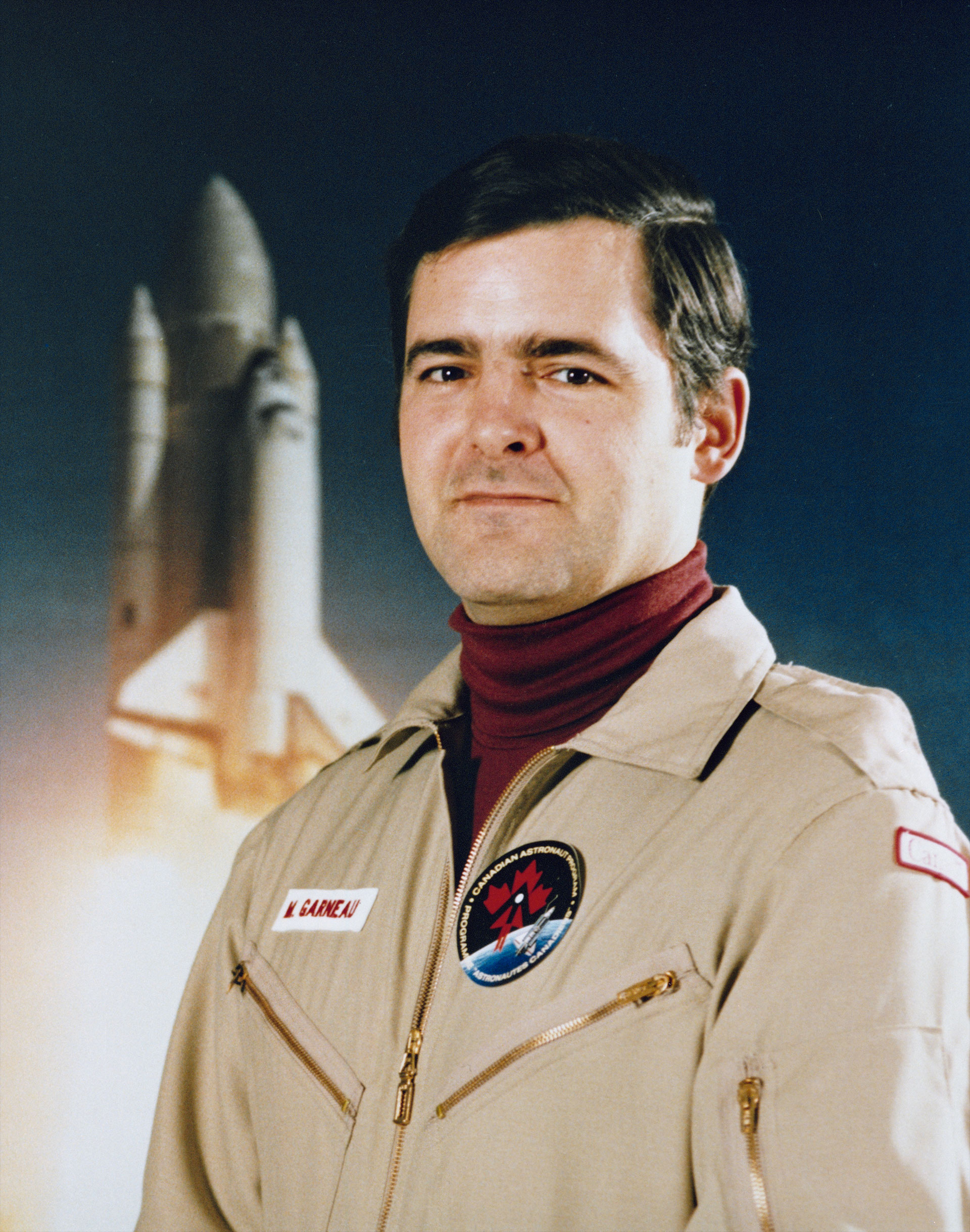 a man poses in front of the image of a space shuttle while wearing a maroon turtleneck shirt and a tan flight suit with patches sewn on its chest.