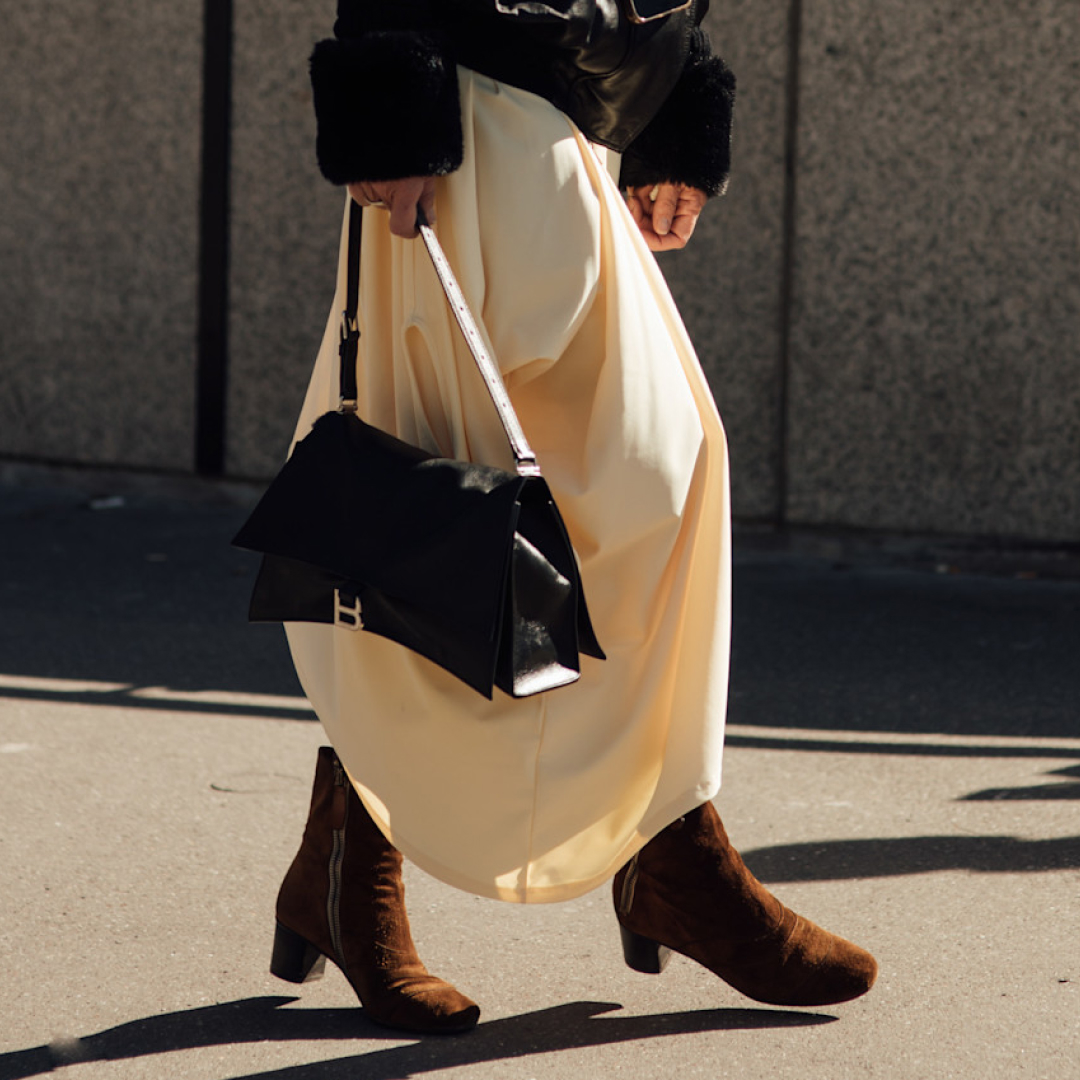 a woman walking on a sidewalk wearing a black fur sleeve jacket, white skirt, brown suede booties, and a black handbag.